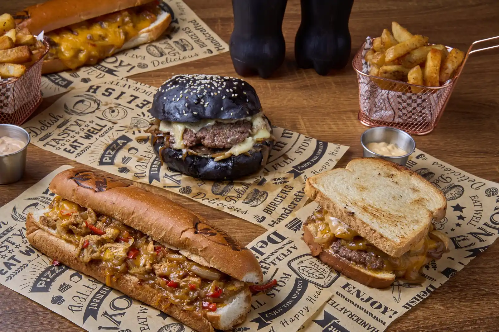 A Phillyzon table: cheesesteak, burger on a black bun, patty melt and fries on kraft paper